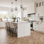 A modern kitchen with herringbone vinyl flooring in a light oak finish, laid in a zigzag pattern beside white cabinets.